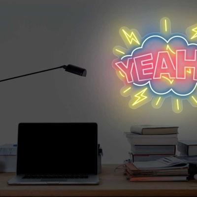 Secretária com portátil, lâmpada e néon colorido com o texto 'YEAH!' na parede