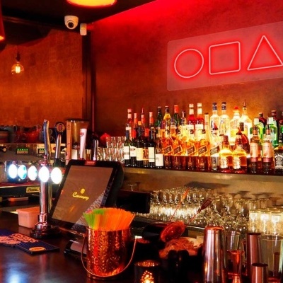 Balcão de bar com garrafas, copos e símbolos geométricos vermelhos na parede