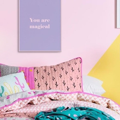 Cama com roupa de cama e almofadas coloridas em quarto com paredes rosa e amarelo, quadros com texto e lâmpada de mesa cinzenta.