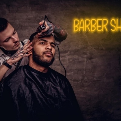 Barbeiro a cortar cabelo a cliente em ambiente com parede cinzenta e letreiro amarelo 'BARBER SHOP'.