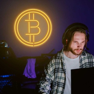 Homem com auriculares mostra símbolo de Bitcoin neon amarelo na parede roxa atrás dele