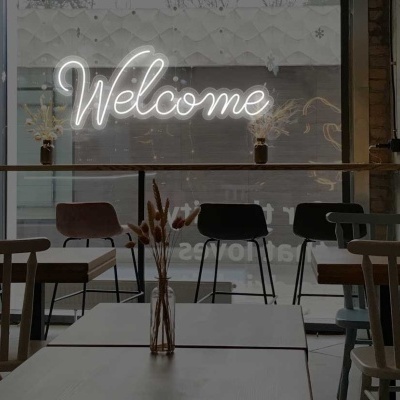 Interior de café com cadeiras variadas, mesas claras e letreiro de neon com a palavra Welcome.