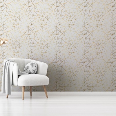 Poltrona cinzenta clara com almofada, manta cinza e candeeiro dourado diante de parede branca com padrão geométrico dourado