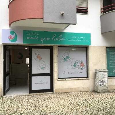 Fachada da clínica mais que bebé com placa verde e branca, janelas decoradas com desenhos de bebés e calçada portuguesa à frente