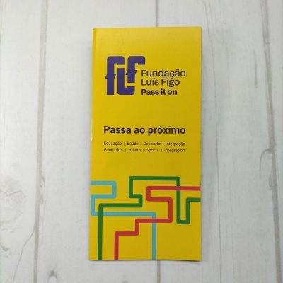 Folheto amarelo da Fundação Luís Figo com texto e linhas coloridas sobre superfície de madeira clara