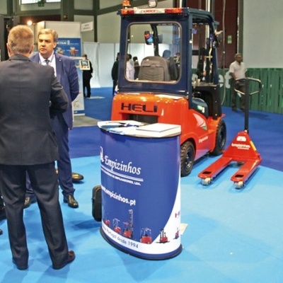 Empilhador industrial HELI laranja e azul num espaço de exposição com três homens e expositor azul escuro