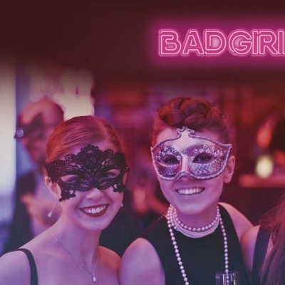 Três mulheres com máscaras decorativas em festa com luzes roxas e texto em néon 'BADGIRLSONLY' ao fundo