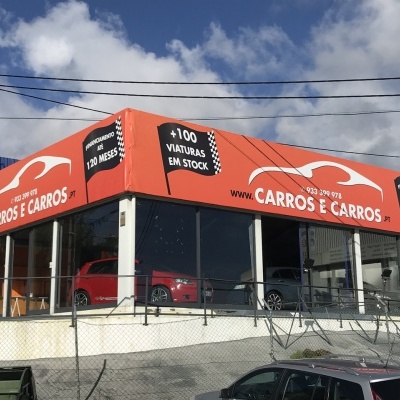 Loja de carros com letreiro laranja e texto branco, carros dentro e fora da loja