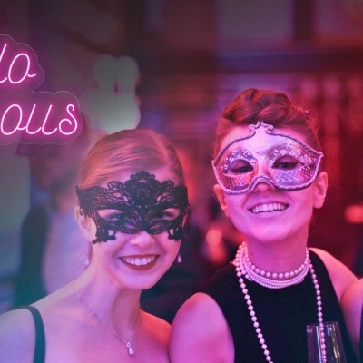 Três mulheres com máscaras de renda em ambiente festivo com letreiro 