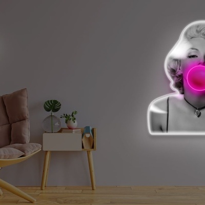 Luminária de parede de neón de uma mulher a soprar chiclete cor-de-rosa na parede cinza ao lado de poltrona bege e mesa branca com plantas.