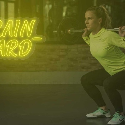Mulher a fazer agachamento com barra numa sala de ginásio com letreiro TRAIN HARD em néon amarelo