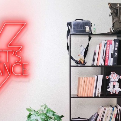 Estante preta com livros e objetos decorativos, luz de néon vermelha com texto LET'S DANCE na parede branca, planta verde