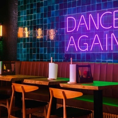 Restaurante/bar com bancos de couro castanho, cadeiras de madeira azuis, mesas de madeira, parede com azulejos verdes e letreiro neon rosa DANCE AGAIN