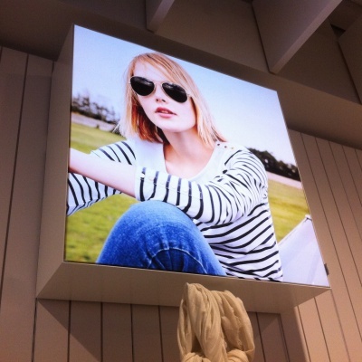 Publicidade de mulher com camisola às riscas e óculos de sol exibida numa moldura iluminada.