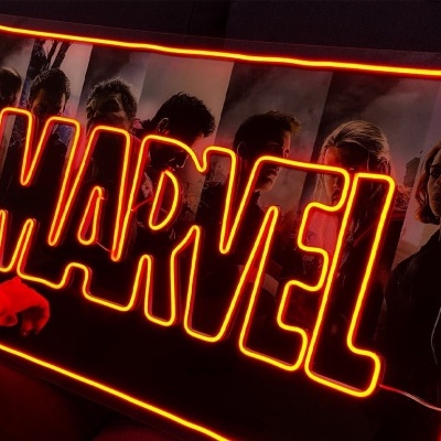 Letreiro luminoso MARVEL em tons de vermelho e amarelo sobre sofá roxo.