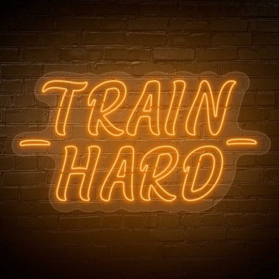Letreiro neon laranja com o texto TRAIN HARD numa parede de tijolos escuros