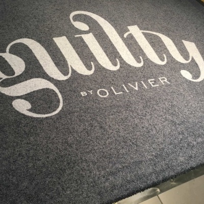 Tapete cinza escuro com texto branco 'guilty BY OLIVIER' no chão