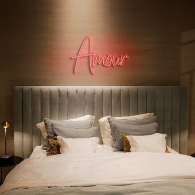Cabeceira de cama cinzenta estofada com roupa branca e luz neon rosa 'Amour' na parede