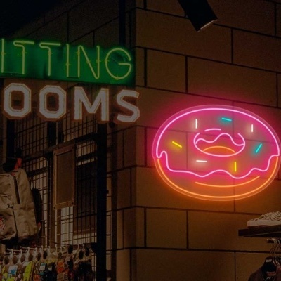 Placas de néon verde FITTING ROOMS e donut rosa numa loja com paredes azulejadas e roupa exposta