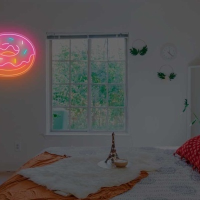 Quarto com cama estofada, almofadas coloridas, manta castanha, plantas e luz néon de donut