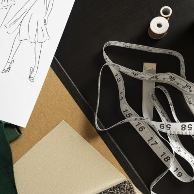 Materiais de design de moda incluindo tecidos, fita métrica, carretel de linha, alfinetes e esboços de vestidos.