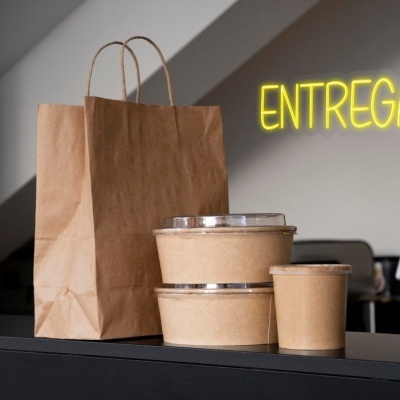 Embalagens para entrega incluindo saco de papel castanho, recipientes de cartão e copo, com letreiro ENTREGAS em néon amarelo ao fundo