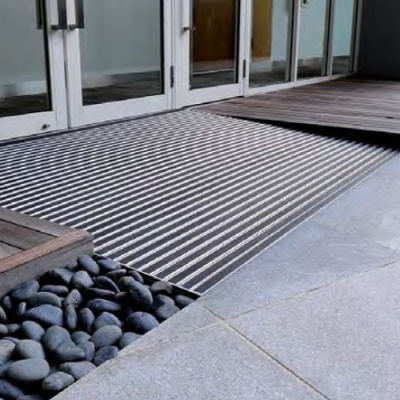 Capacho metálico antiderrapante exterior em frente a portas de vidro, com pedras pretas e piso de madeira e azulejos.