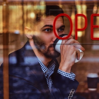 Homem a beber café num copo descartável dentro de uma cafetaria com a palavra OPEN na janela