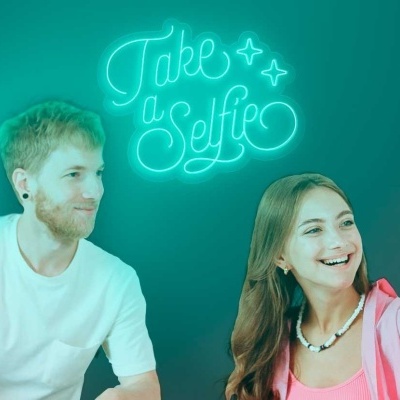 Dois jovens tiram selfie com luz verde e texto 'Take a Selfie' ao fundo