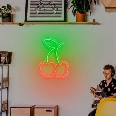 Parede branca com letreiro de néon em forma de cerejas, prateleira, estante com embalagens e pessoa lendo sentada.