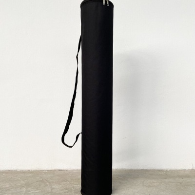 Bolsa cilíndrica preta com fecho e alça, apoiada contra parede branca