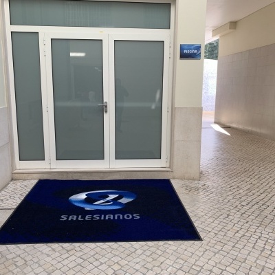 Tapete azul com logotipo e texto 'SALESIANOS' num chão de calçada portuguesa junto a porta de vidro
