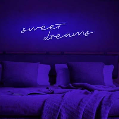 Cama de casal com almofadas e manta, letreiro de néon azul com texto 'sweet dreams' na parede roxa.