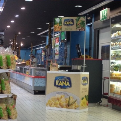 Caixa promocional de massas Rana no corredor de supermercado com sacos de massa à esquerda e frigorífico com produtos à direita.