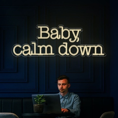 Lâmpada de neon 'Baby calm down' numa parede azul escuro com homem a trabalhar no portátil.