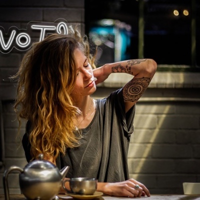 Mulher sentada à mesa com chaleira e xícara, texto 'WHY NOT' iluminado na parede