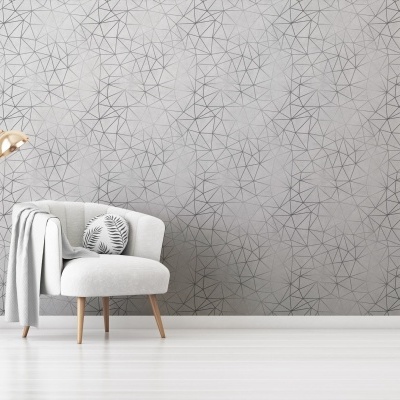 Poltrona cinzenta clara com almofada estampada e candeeiro dourado ao lado sobre fundo de parede geométrica cinzenta.