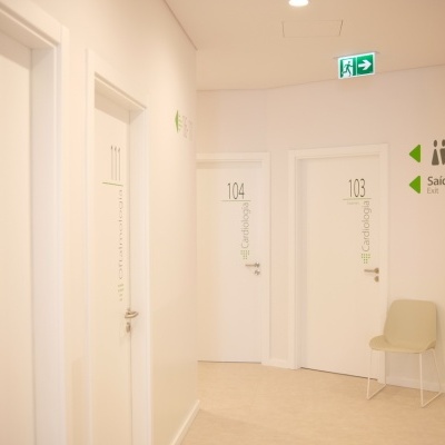 Corredor branco com portas numeradas e sinais verdes de saída e sanitários, com uma cadeira bege encostada na parede.