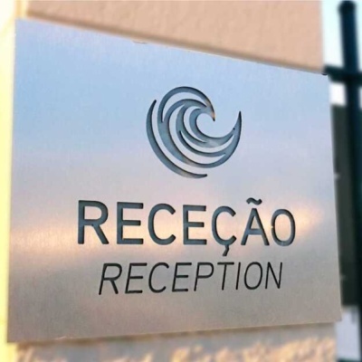 Placa metálica prateada com texto 'RECEÇÃO' e 'RECEPTION' e logotipo circular