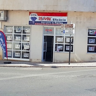 Fachada da agência imobiliária RE/MAX Eficácia com listagens de imóveis nas janelas e bandeiras publicitárias