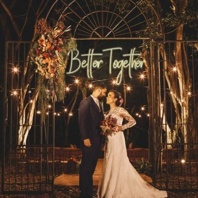 Noivos sob arco metálico iluminado com a frase 'Better Together' em cenário noturno festivo