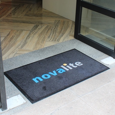 Tapete preto com texto 'novalite' na entrada de edifício, chão azulejos cinzentos e madeira