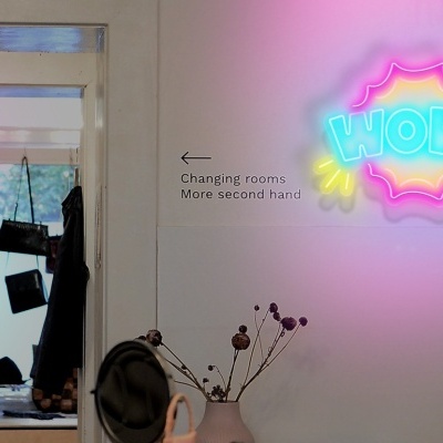 Casaco preto pendurado e letreiro neon 'WOW!' em parede branca com sinalização para provadores.