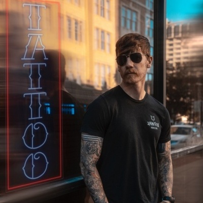 Homem tatuado de óculos escuros e t-shirt preta junto a janela com letreiro 'TATTOO'