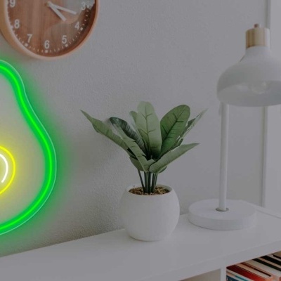 Neon verde e amarelo em forma de abacate numa parede junto a relógio, planta em vaso e candeeiro sobre prateleira branca