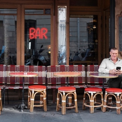 Cadeiras e bancos em esplanada de café com rapaz sentado a olhar para telemóvel