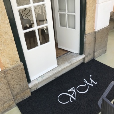 Tapete de entrada preto com letras brancas 'AM' junto a porta de vidro e madeira branca