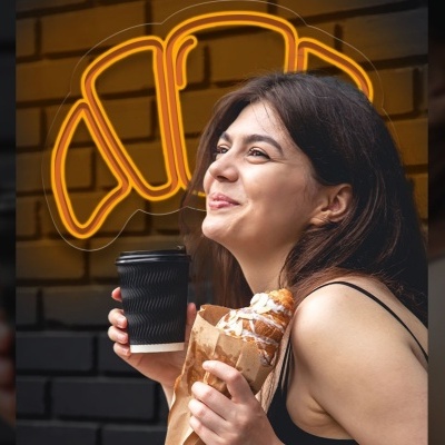 Mulher segurando copo preto e saco com croissants em frente a neon de croissant amarelo