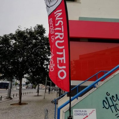 Bandeira publicitária de escola de condução junto a escada e placa informativa numa rua com árvores