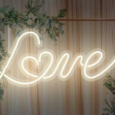 Letreiro de néon branco a dizer 'Love' com coração, decorado com ramos verdes e cortinas bege ao fundo